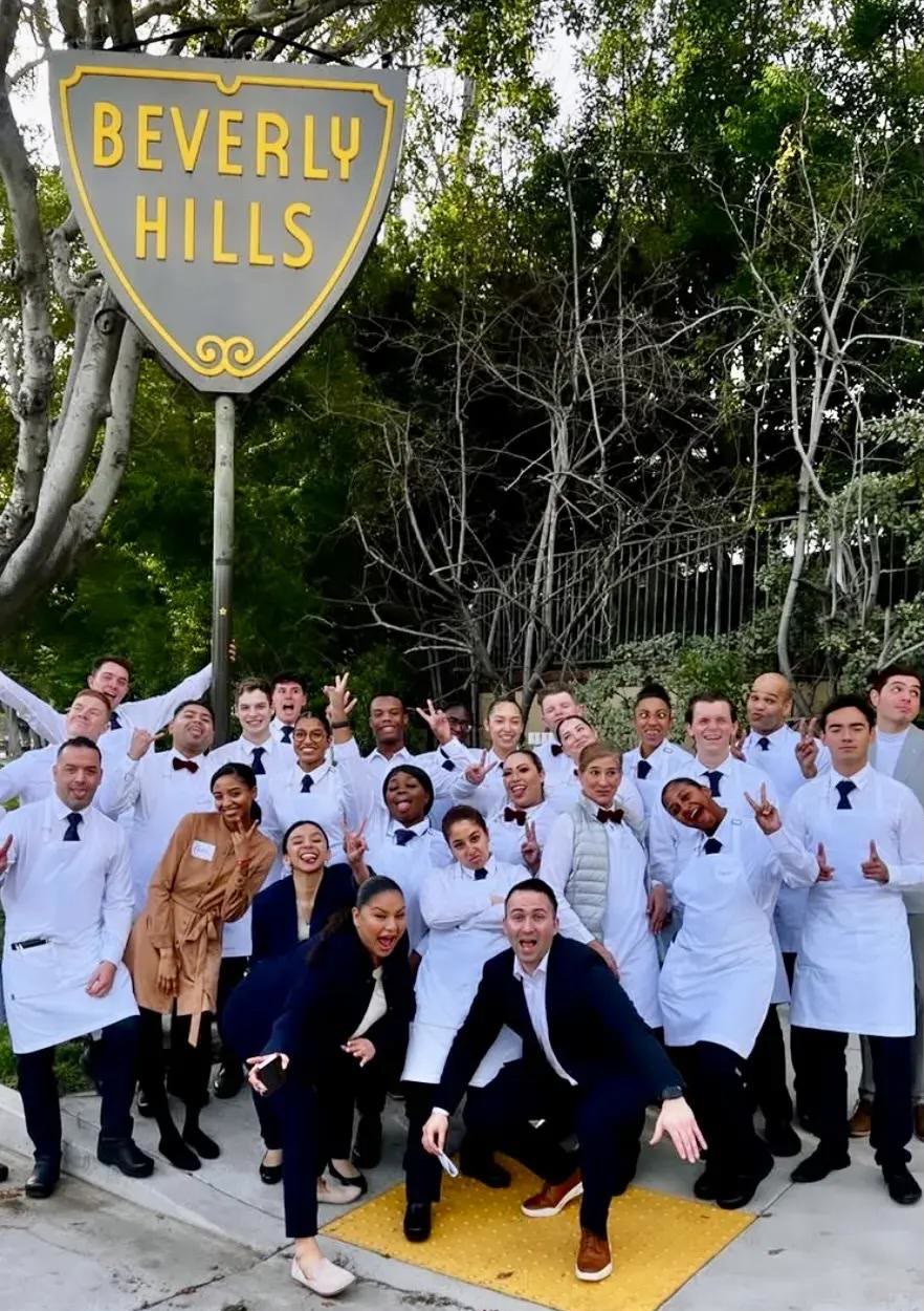 An opening team in white service jackets and aprons, arms raised, gathered under the gold-lettered Beverly Hills city shield sign