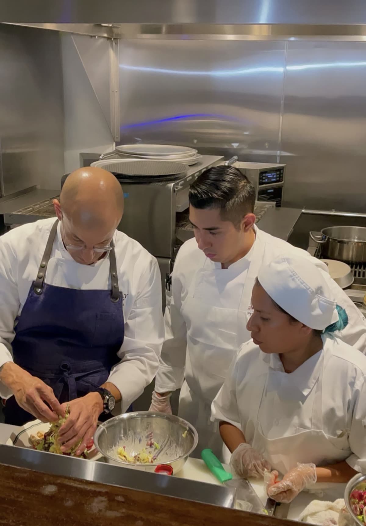 A chef in a blue apron working a dish through with two cooks at a kitchen station