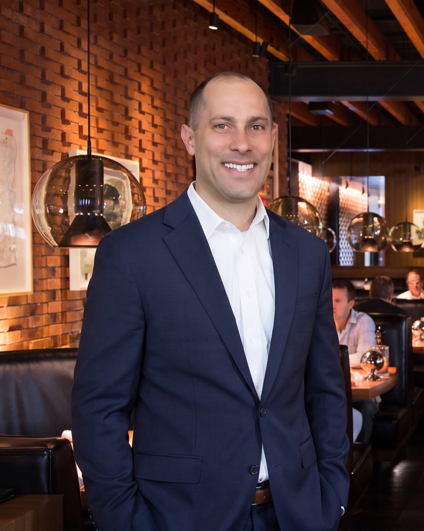Jon Peck in a navy suit, standing in a brick-walled restaurant dining room with the open kitchen behind him