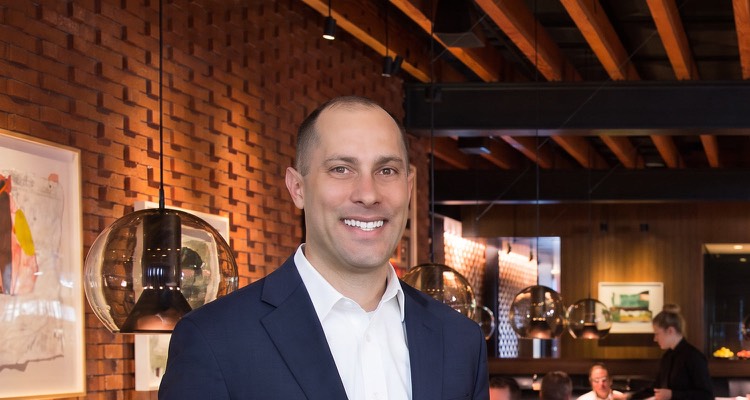 Jon Peck smiling in a brick-walled restaurant dining room, pendant lights behind him