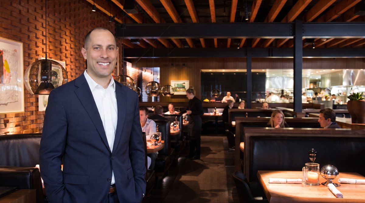 Jon Peck standing in a brick-walled restaurant dining room, with booths, pendant lights and the open kitchen behind him
