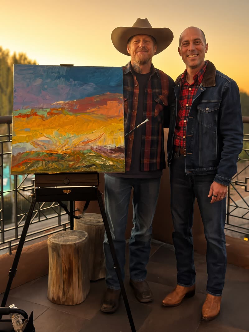 Jon Peck, smiling in a denim jacket and plaid shirt, on a rooftop at sunset beside a painter in a cowboy hat and his landscape canvas on an easel