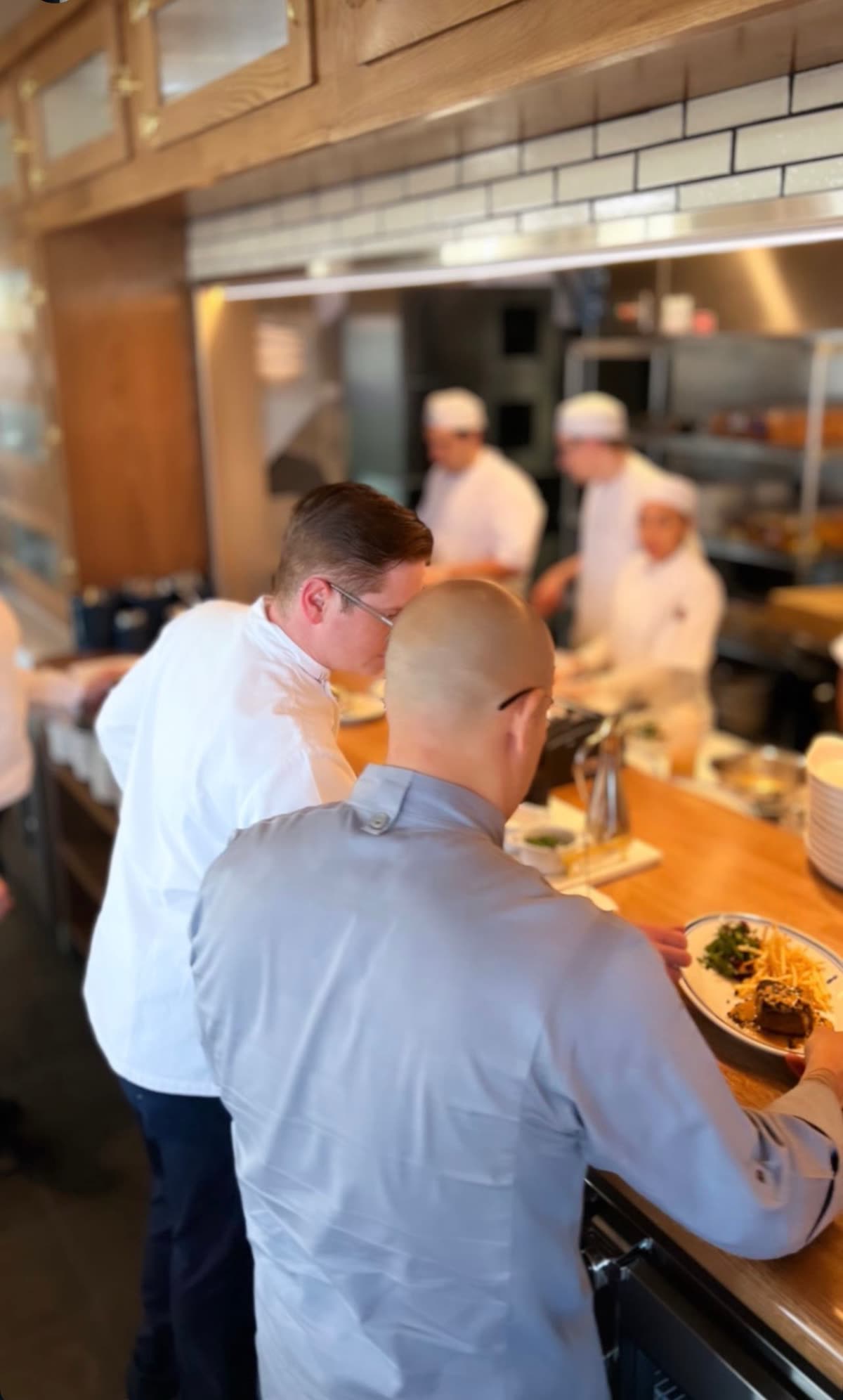 Two chefs checking a plate of steak frites at the pass while cooks work the line behind them